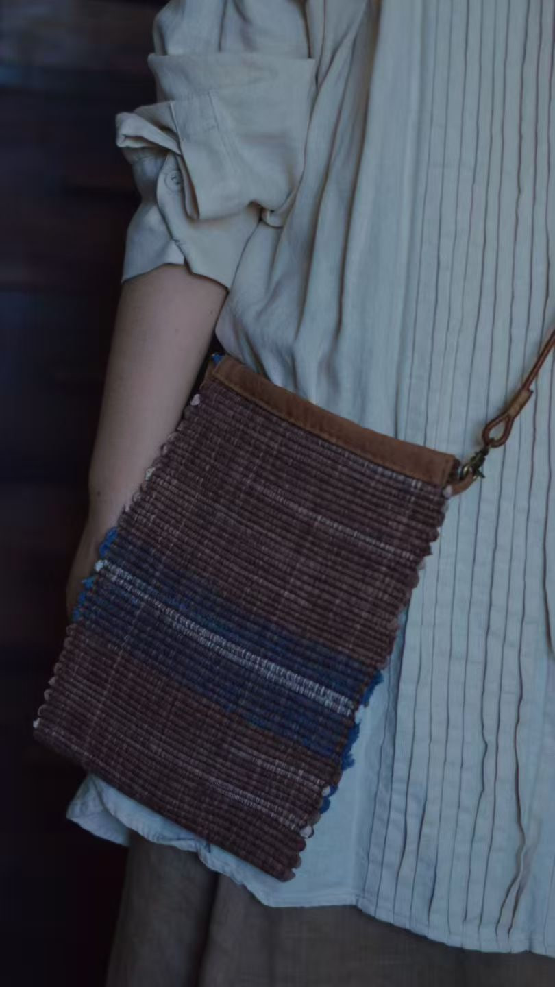 A person in a pleated pale shirt wearing a brown woven crossbody bag with blue stripes.