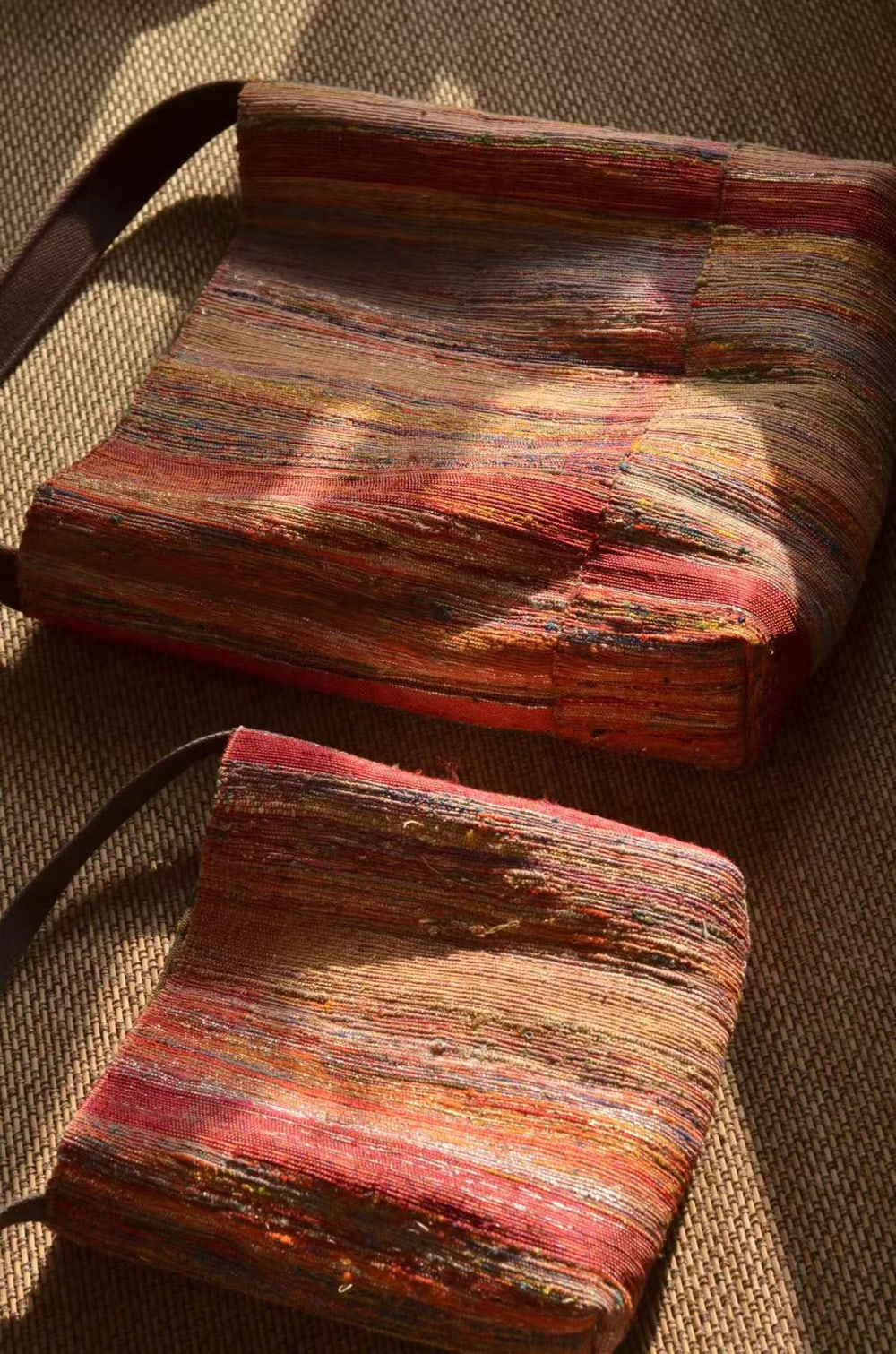 Two woven shoulder bags in red, orange and pink stripes lying on a burlap surface.
