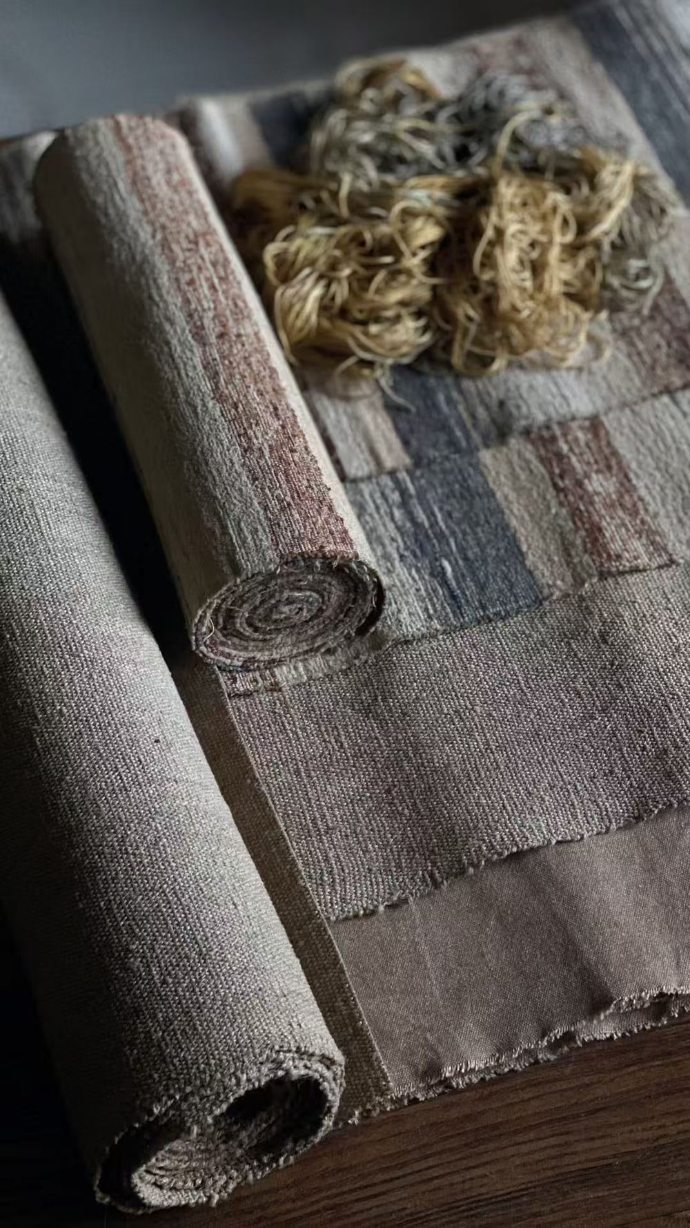Striped hand-woven cloth rolled beside a loose pile of raw fibre on a dark wooden surface.