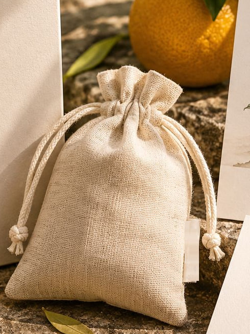 Concept render: a linen drawstring sachet among citrus leaves and blossom, with an orange behind.