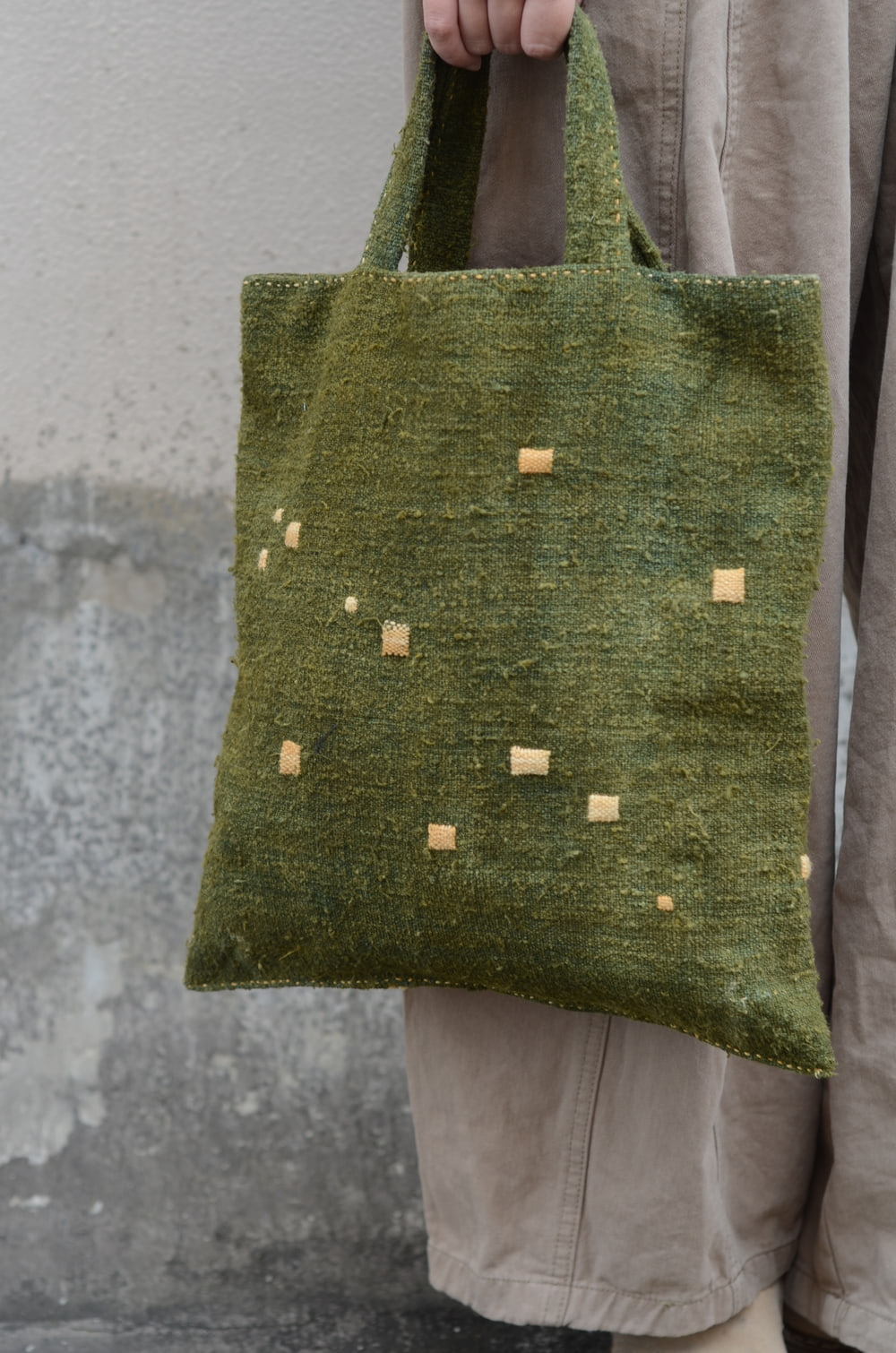 A deep green textured tote bag held by one of its two handles against a pale stone wall, with small light-coloured square patches scattered across the front.